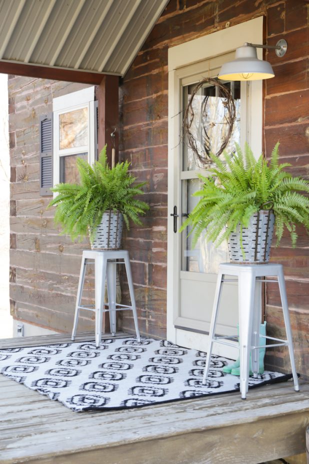 Rustic Log Home With a Farmhouse Decor Porch Theme, Olive Buckets, a Handmade Grapevine Wreath, Galvanized Stools, and a Black and White Reversible Outdoor Rug