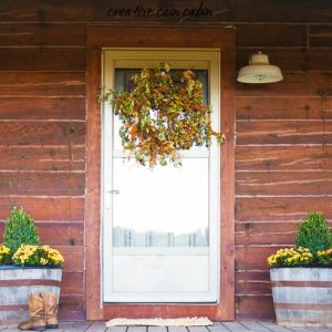 Rustic Front Porch of a Log Cabin Decorated For Fall Using Whiskey Barrels, Boxwoods, Mums, and Bittersweet