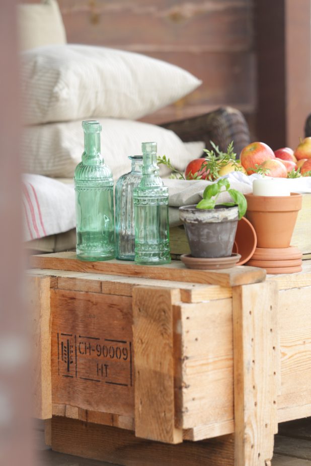 Rustic fall porch decor with apples, a shipping crate, sea glass green bottles, herbs, terra cotta pots, and ticking pillows